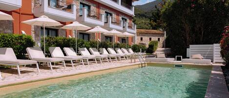 A poolside area with lounge chairs and umbrellas, a stone building with balconies, and a mountain in the background.