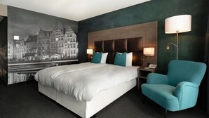 A hotel room with a large bed, a nightstand, a chair, and a wall-mounted lamp. - Van der Valk Hotel Nazareth - Gent (Nazareth)