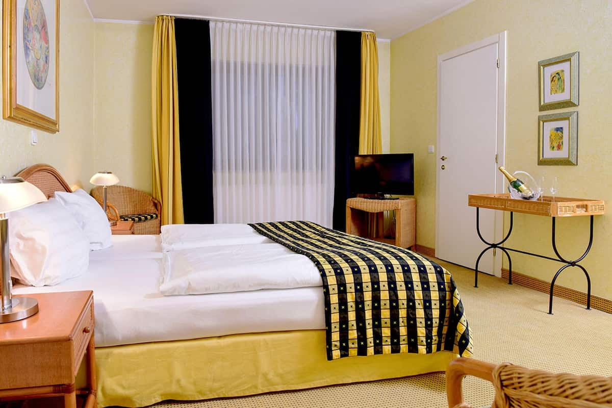 Habitación de hotel con cama, televisor, escritorio con una botella y vasos, una silla y ventana con cortinas.