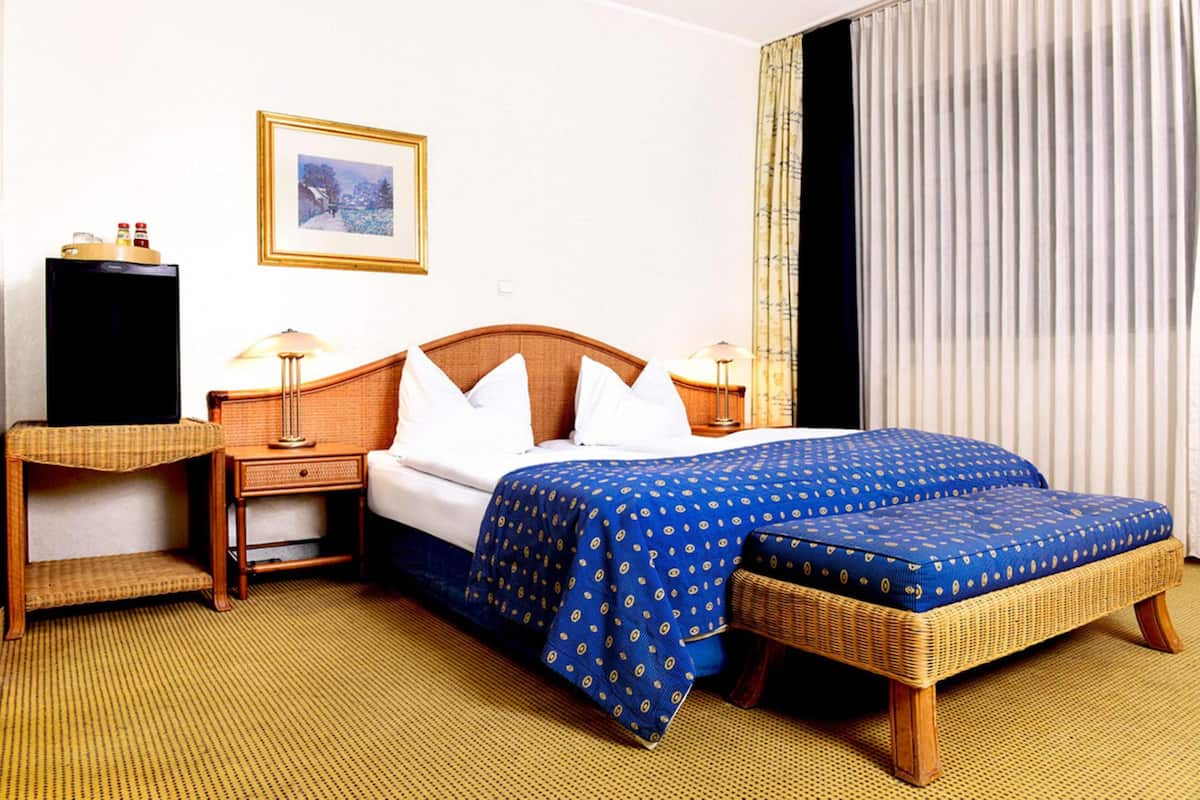Una habitación de hotel con una cama, dos mesitas de noche, una lámpara, un cuadro en la pared y una ventana con cortinas.