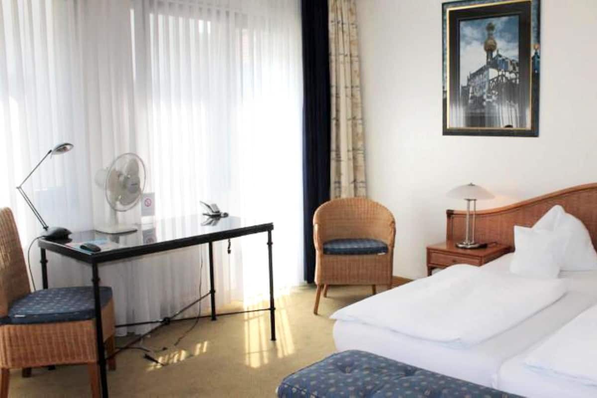 Habitación de hotel con cama, escritorio, silla, lámpara de pie y un cuadro en la pared.