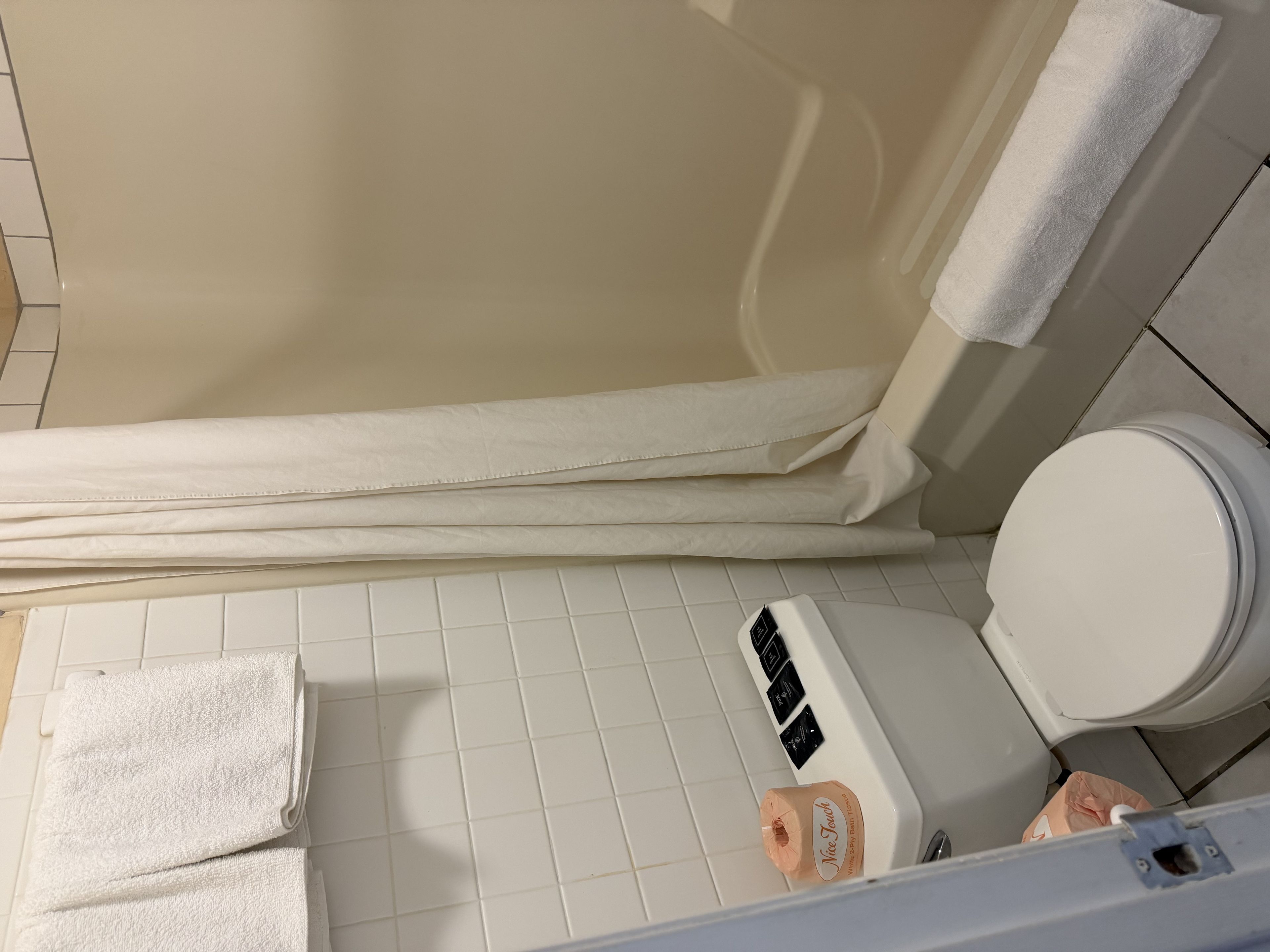A bathroom with a white toilet, a white bathtub, and a white shower curtain. There are two white towels stacked on the floor.