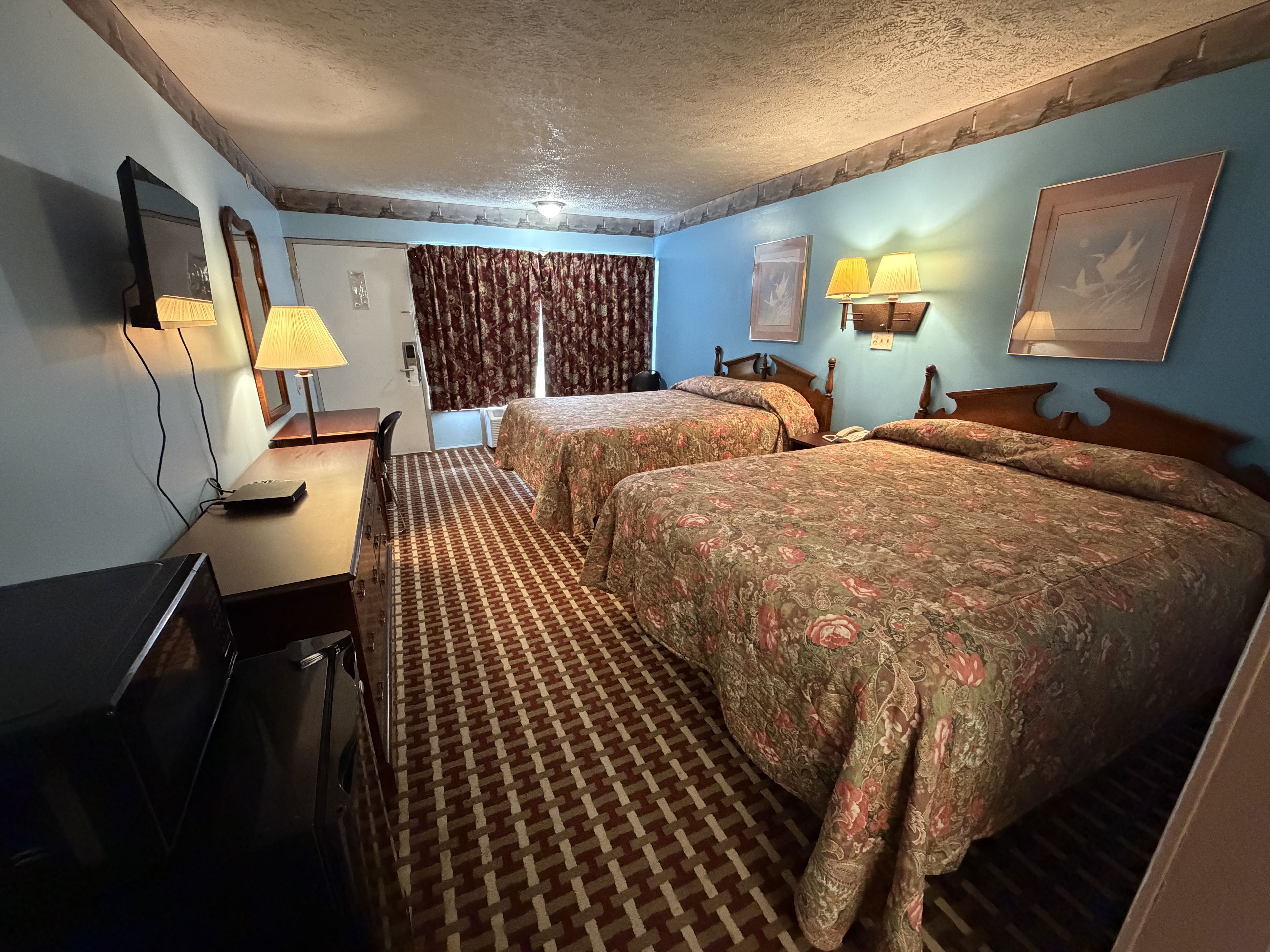 A hotel room with two beds, a desk, a lamp, and a painting on the wall.