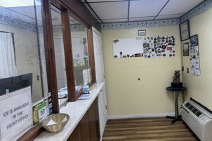 Uma área de recepção com balcão, mural de avisos, pequena mesa com aquecedor e sinalização indicando disponibilidade de gelo.