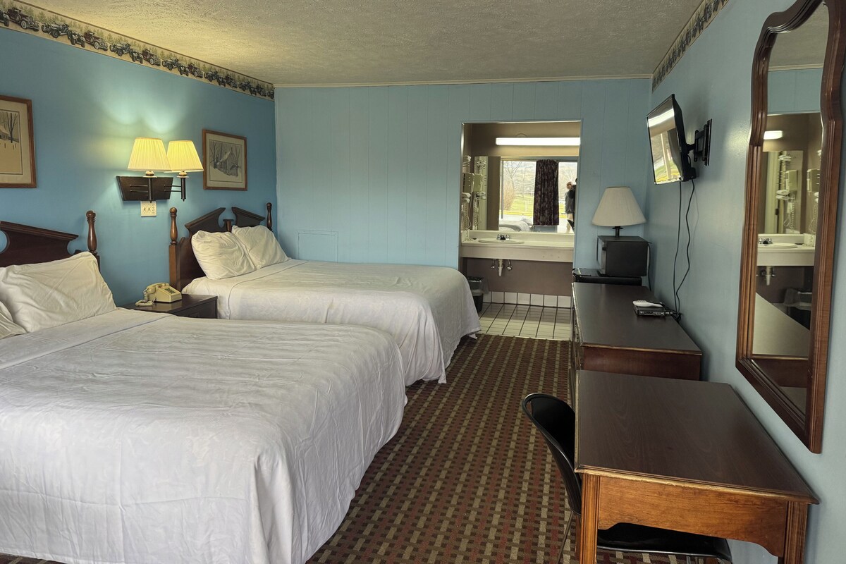 A hotel room with two beds, a desk, a chair, a mirror, a TV, and a bathroom visible through an open door.