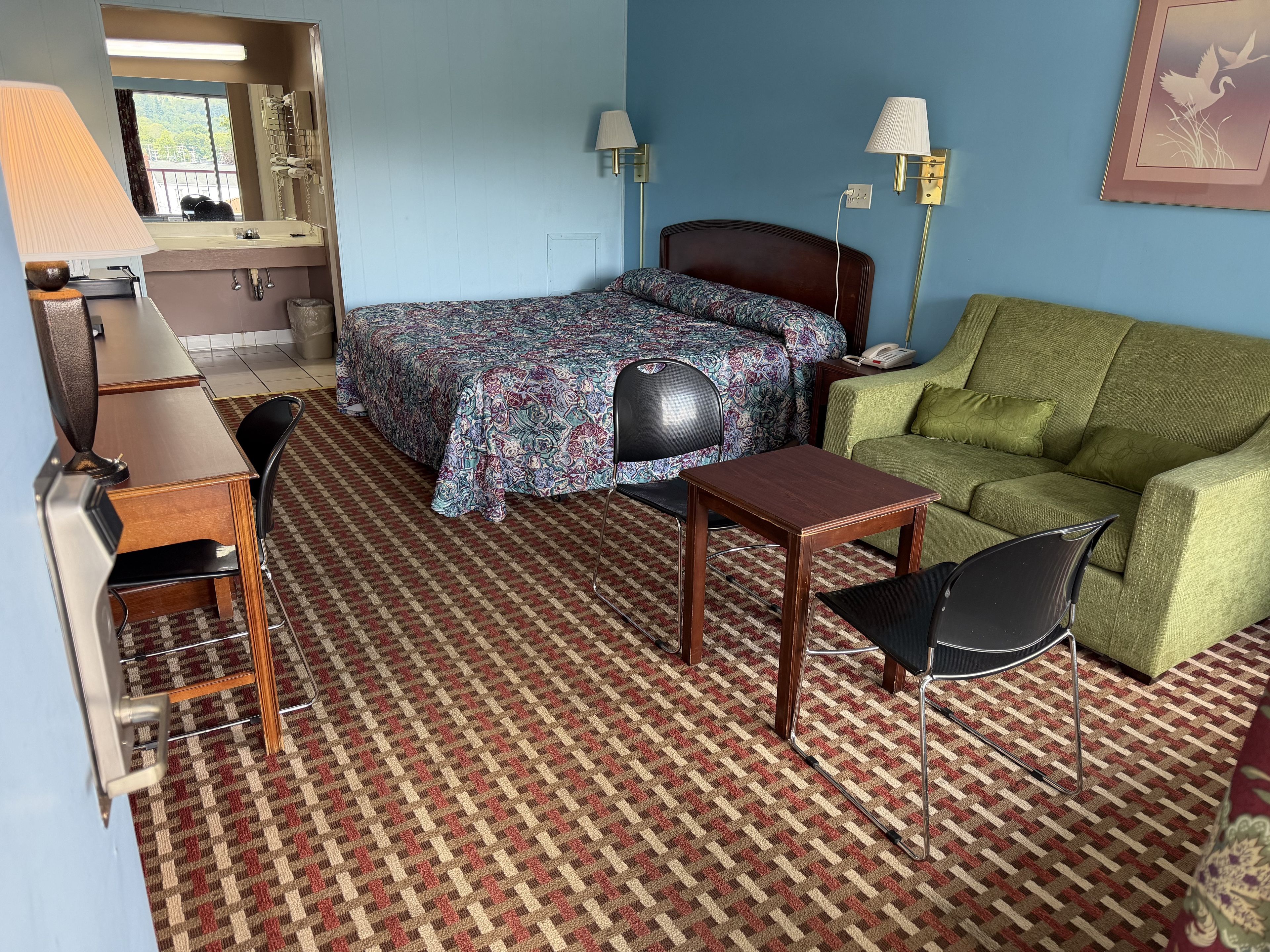 Ett hotellrum med en säng, en grön soffa, ett litet bord, en stol, ett skrivbord och en lampa.