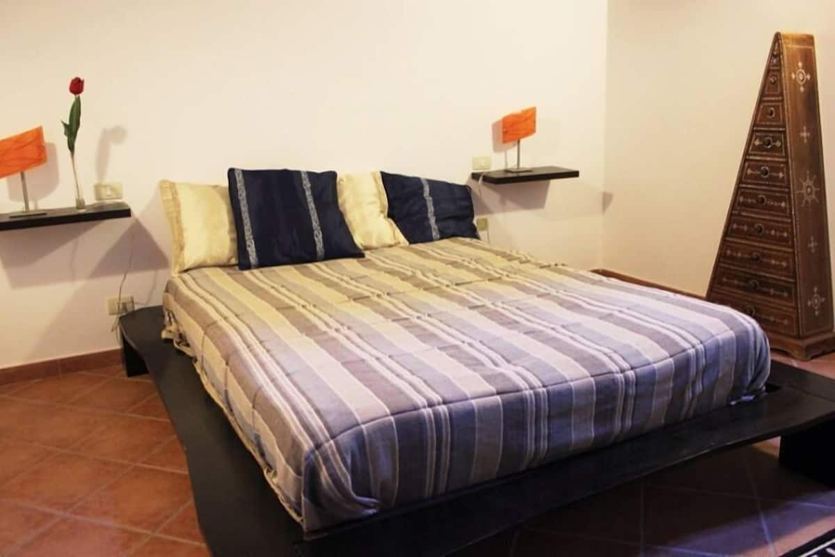 A bed with a striped bedspread, two bedside tables with lamps, a small shelf with a rose, and a wooden cabinet.