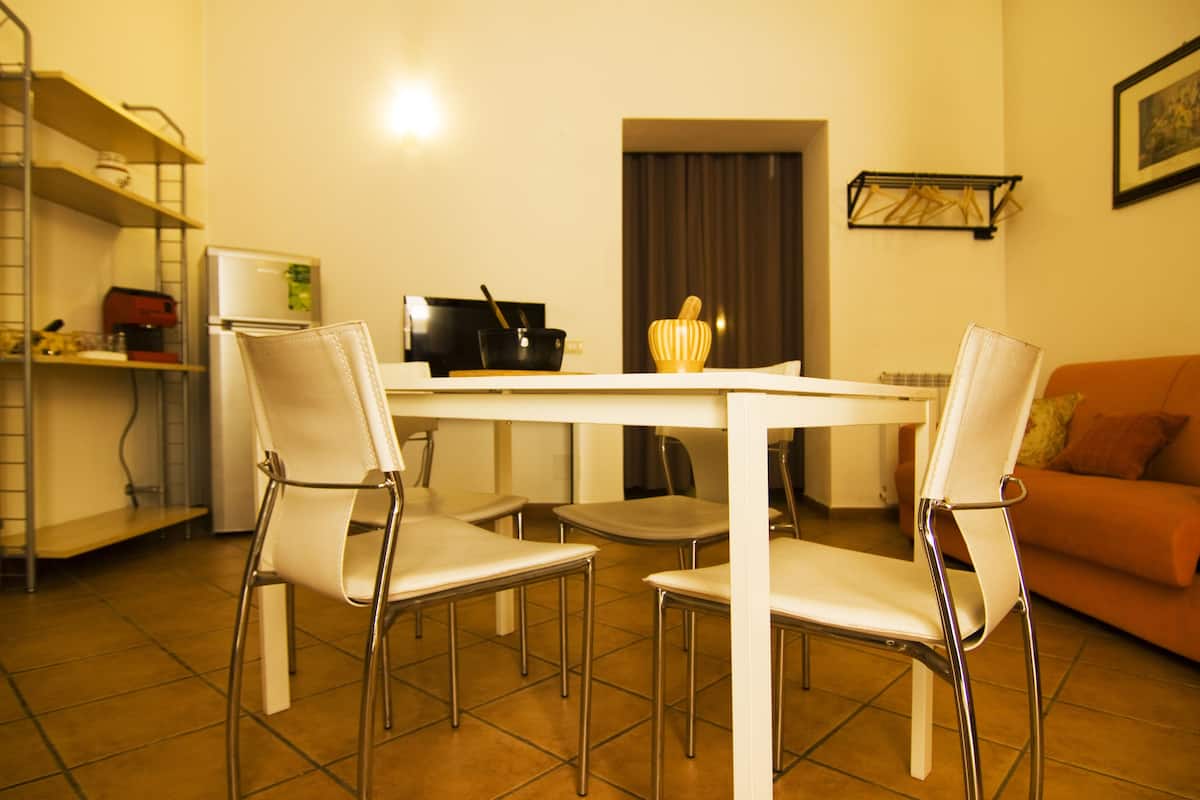 A dining area with a white table and four white chairs, a kitchen area with a refrigerator and a stove, and an orange couch in the background.