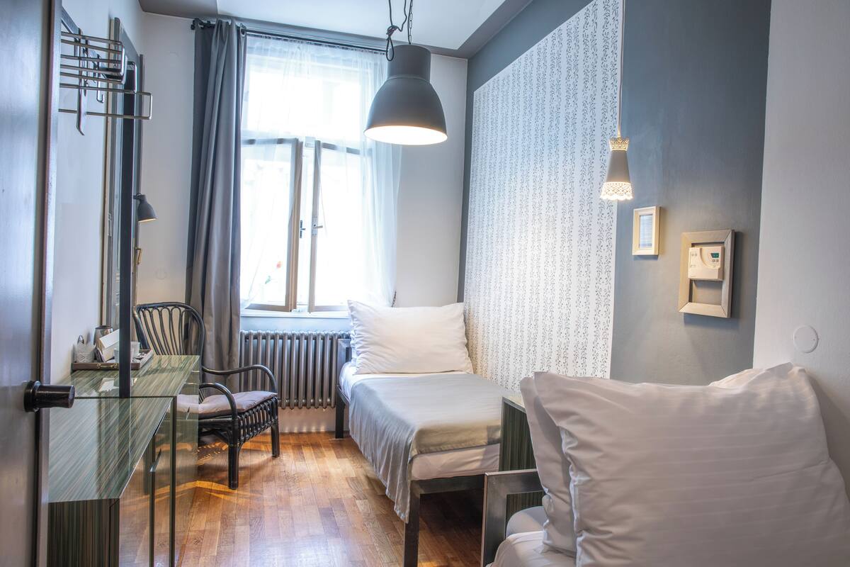 Een kleine slaapkamer met een bed, twee stoelen, een tafeltje en een raam met gordijnen.