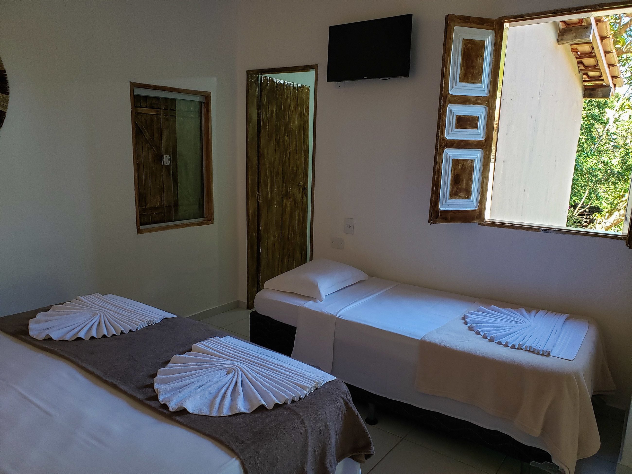 A room with two beds, each with a neatly folded white towel and a brown blanket. A flat-screen TV is mounted on the wall above the beds.