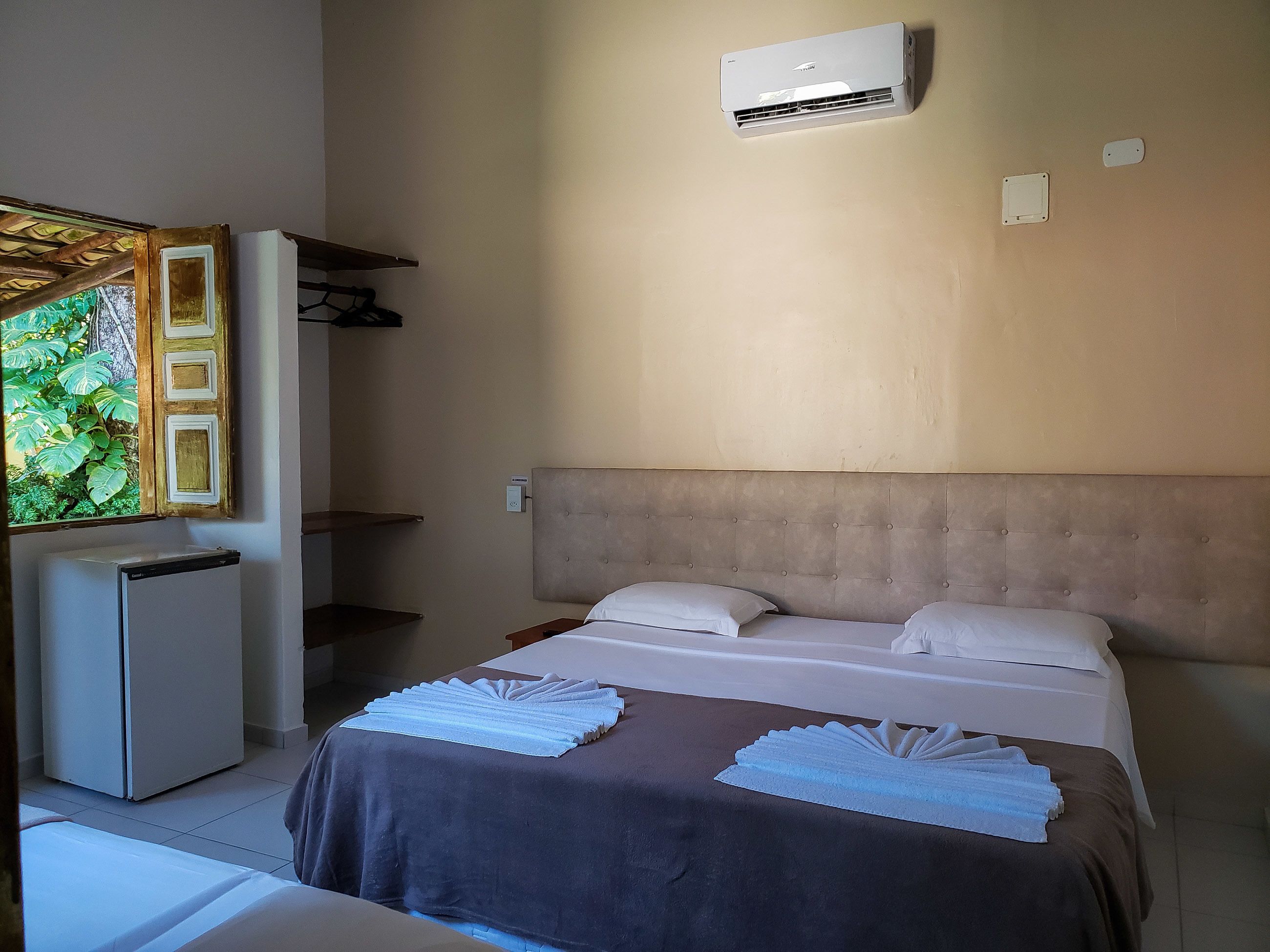 A hotel room with a bed, a small refrigerator, an air conditioning unit, and a window with a view of greenery.