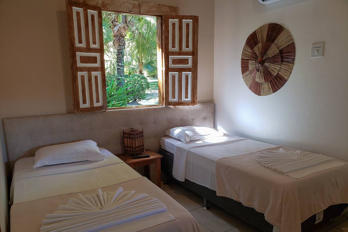 A room with two beds, a window with wooden shutters, a wall-mounted fan, and a view of greenery outside.