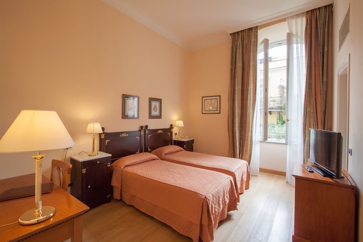 A hotel room with two beds, a desk with a lamp, a TV, and a window with curtains.