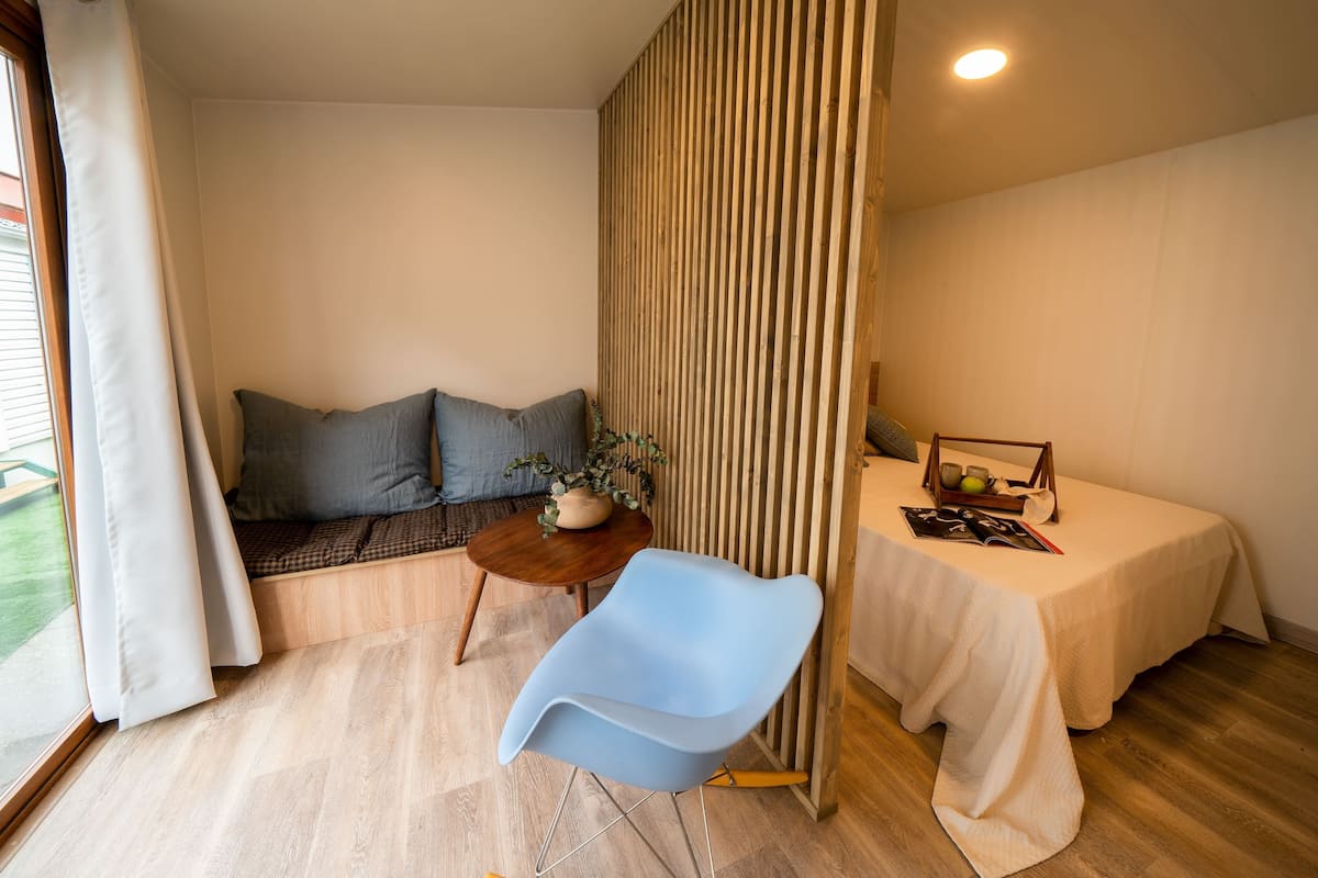 Quarto com cama, uma mesinha com uma planta, uma cadeira azul e uma janela com cortinas.