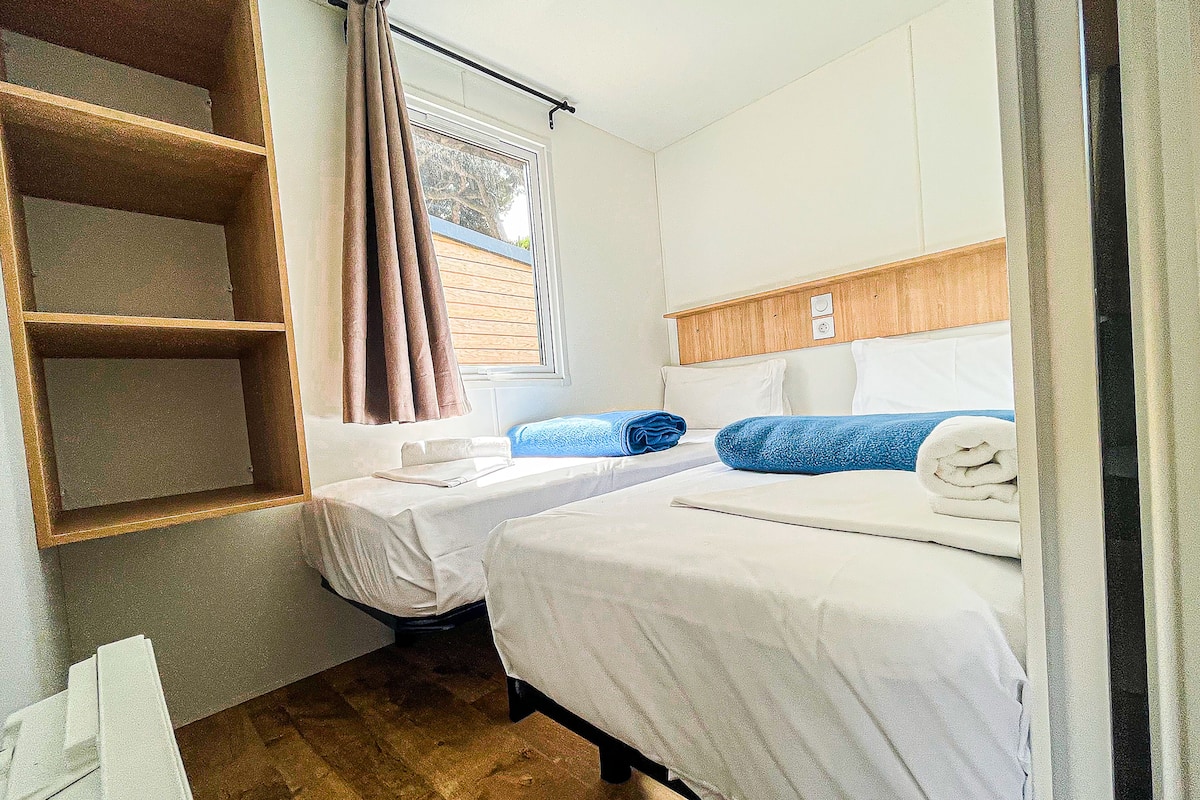Quarto de hotel pequeno e compacto, com duas camas de solteiro, cabeceira de madeira, janela com cortinas e prateleira embutida.