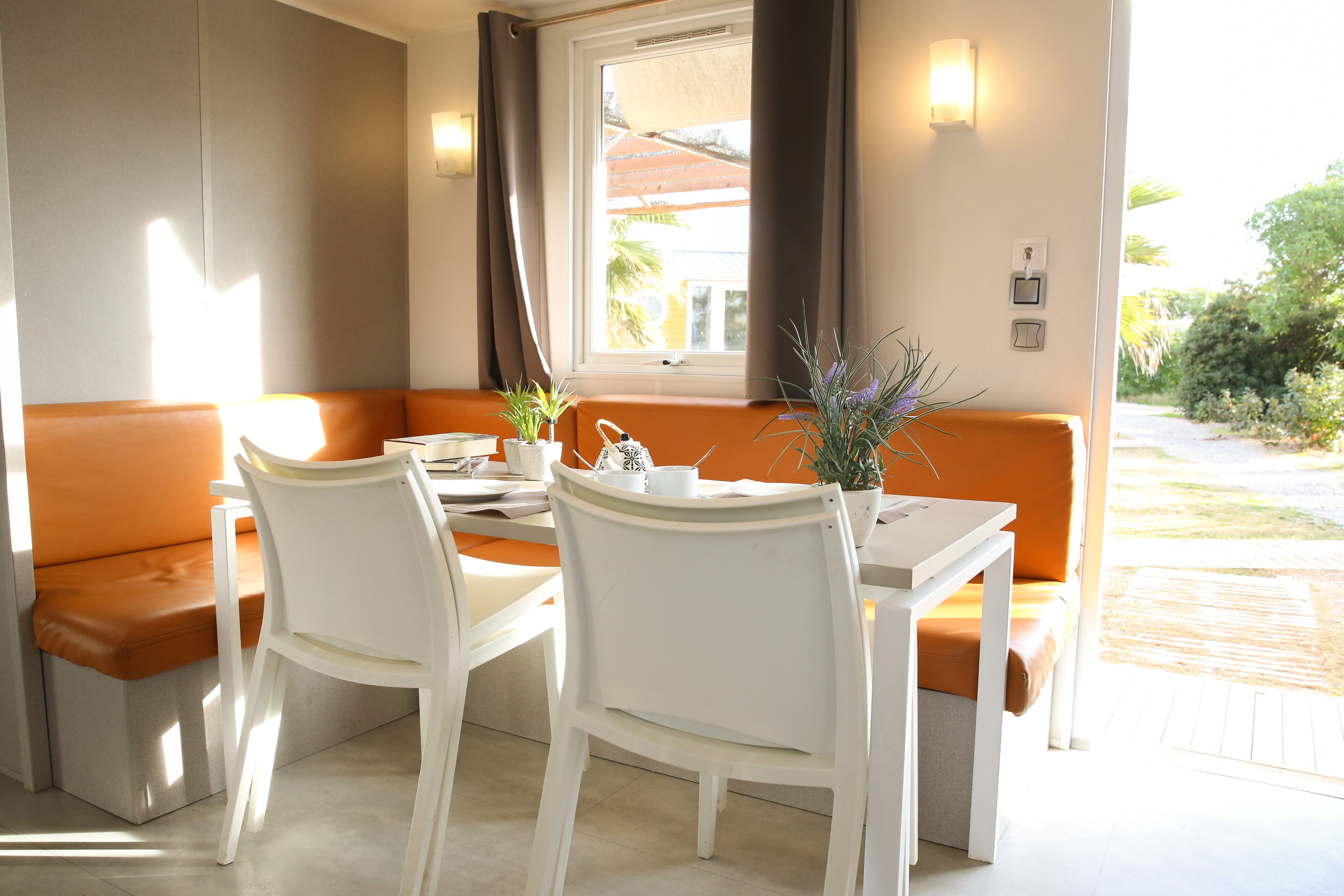 A dining area with a white table, two white chairs, an orange bench, and a window with curtains.