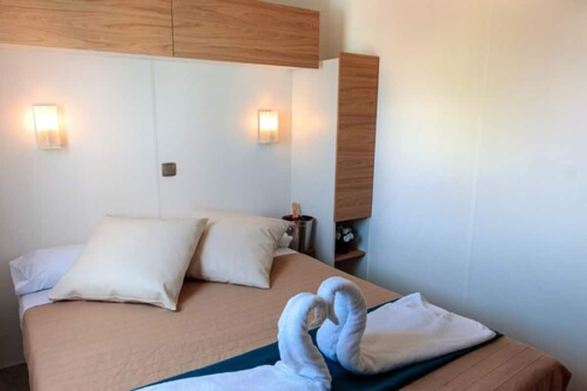 Quarto de hotel com uma cama, duas almofadas, uma mesinha de cabeceira e luminárias fixadas na parede.