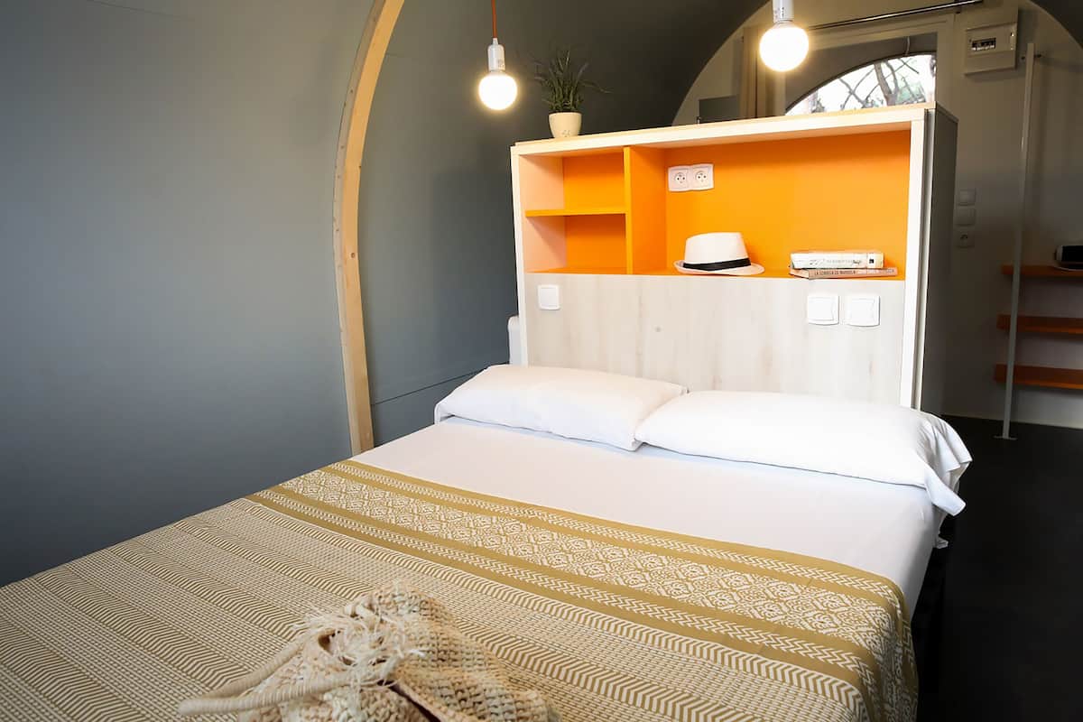 Uma cama com colcha estampada, travesseiro branco e cabeceira branca. Há uma estante acima da cama com uma planta e um chapéu.