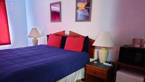 A neatly made bed with a blue cover, two bedside lamps, a small wooden nightstand, a television, and two framed pictures on the wall. - Model Towne Inn (Gwinn)