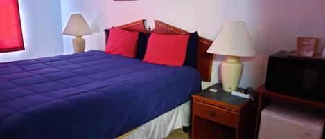 A neatly made bed with a blue cover, two bedside lamps, a small wooden nightstand, a television, and two framed pictures on the wall.