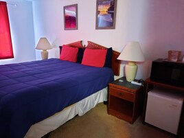 Uma cama bem arrumada com cobre azul, duas luminárias de cabeceira, uma pequena mesa de cabeceira de madeira, uma televisão e duas fotos emolduradas na parede.