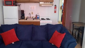 A compact living space with a blue sofa, two red cushions, a white refrigerator, a coffee maker, a sink, a stove, and a fire extinguisher. - Model Towne Inn (Gwinn)