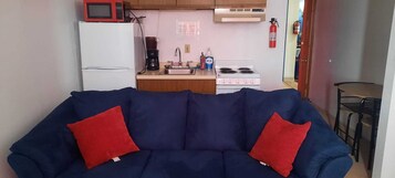 Um espaço compacto com um sofá azul, duas almofadas vermelhas, um frigorífico branco, uma máquina de café, uma pia, um fogão e um extintor de incêndio.