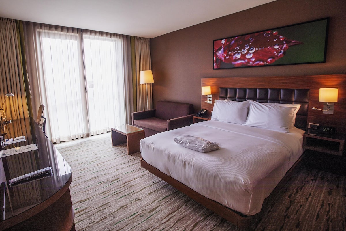 Quarto de hotel com uma cama grande, uma escrivaninha, uma cadeira, uma luminária de mesa, uma mesa de cabeceira e um quadro na parede.