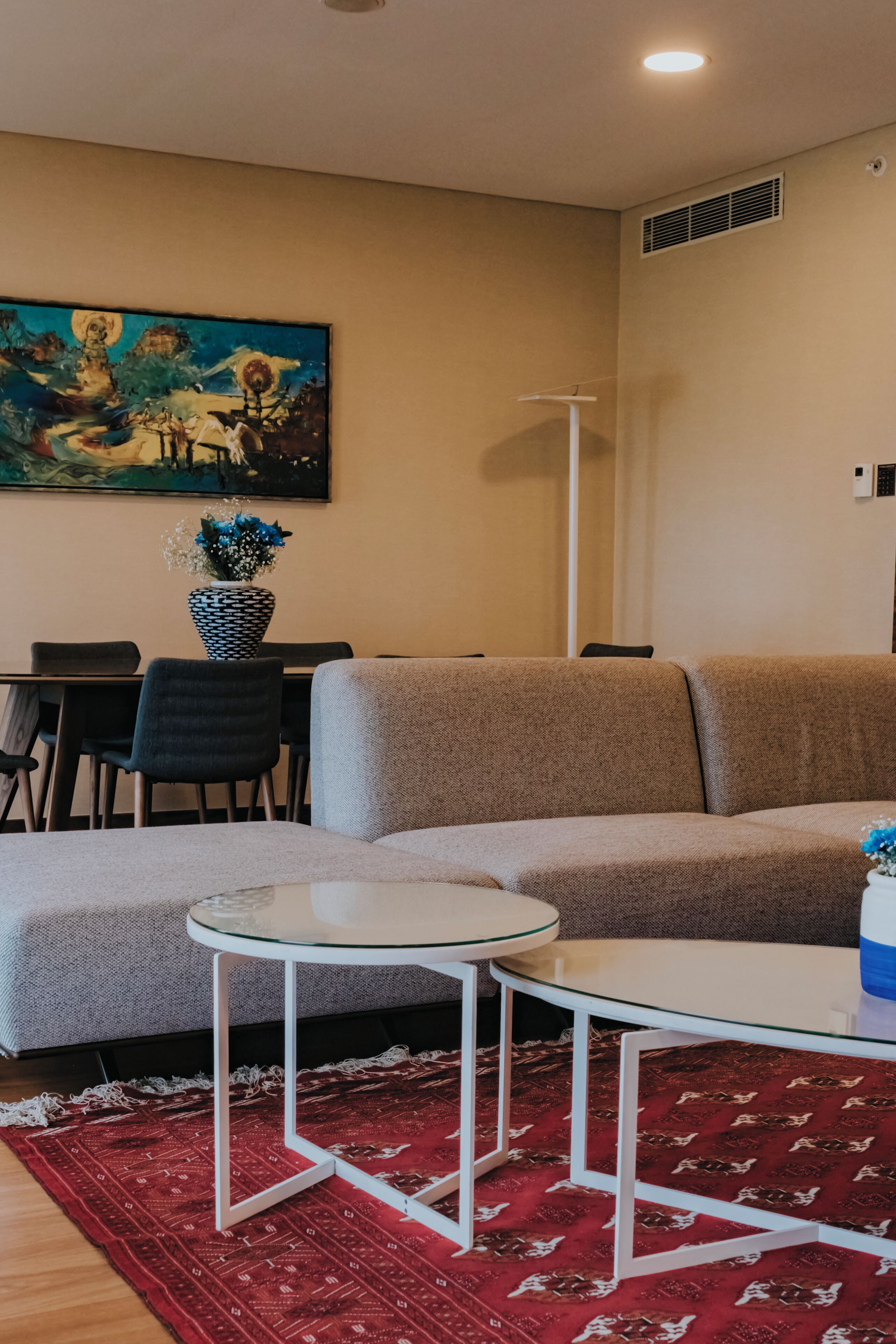 A living room with a sofa, two glass-top tables, a red patterned rug, a painting on the wall, and a dining area with chairs and a table.