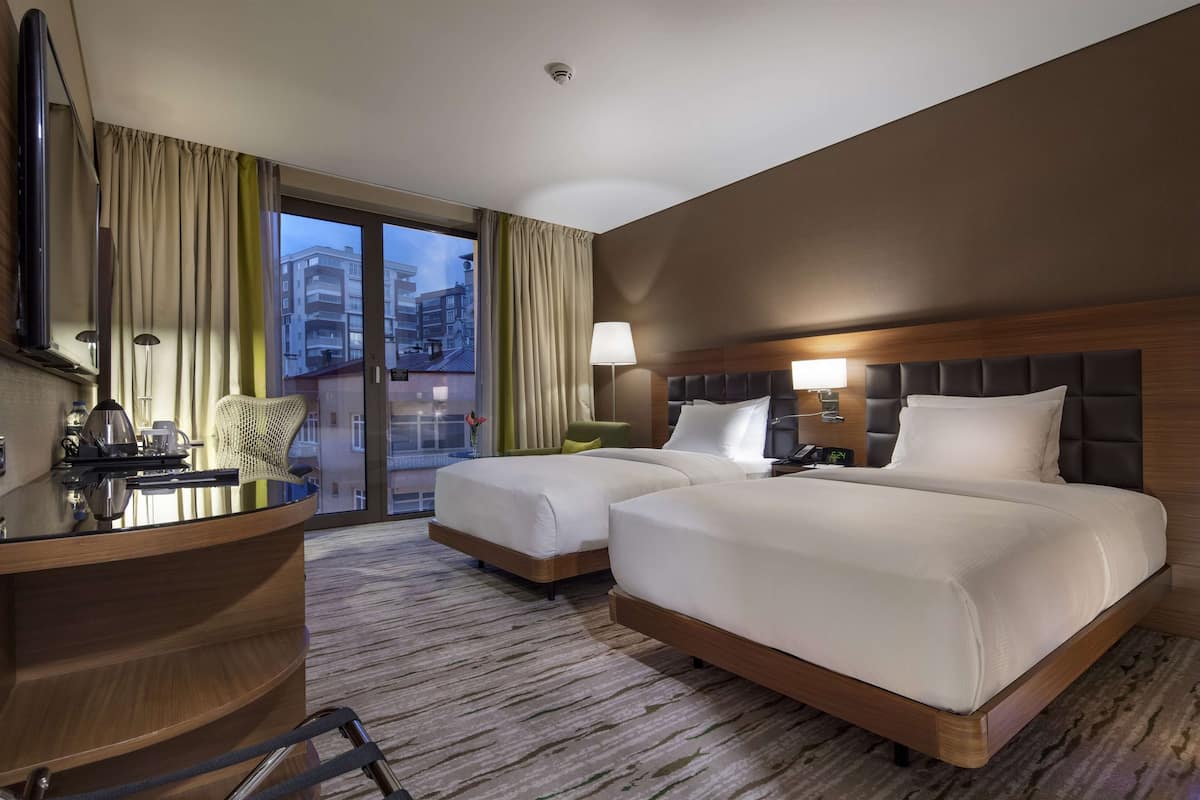 Quarto de hotel com duas camas, uma escrivaninha com cadeira, uma janela grande com cortinas e vista para edifícios.