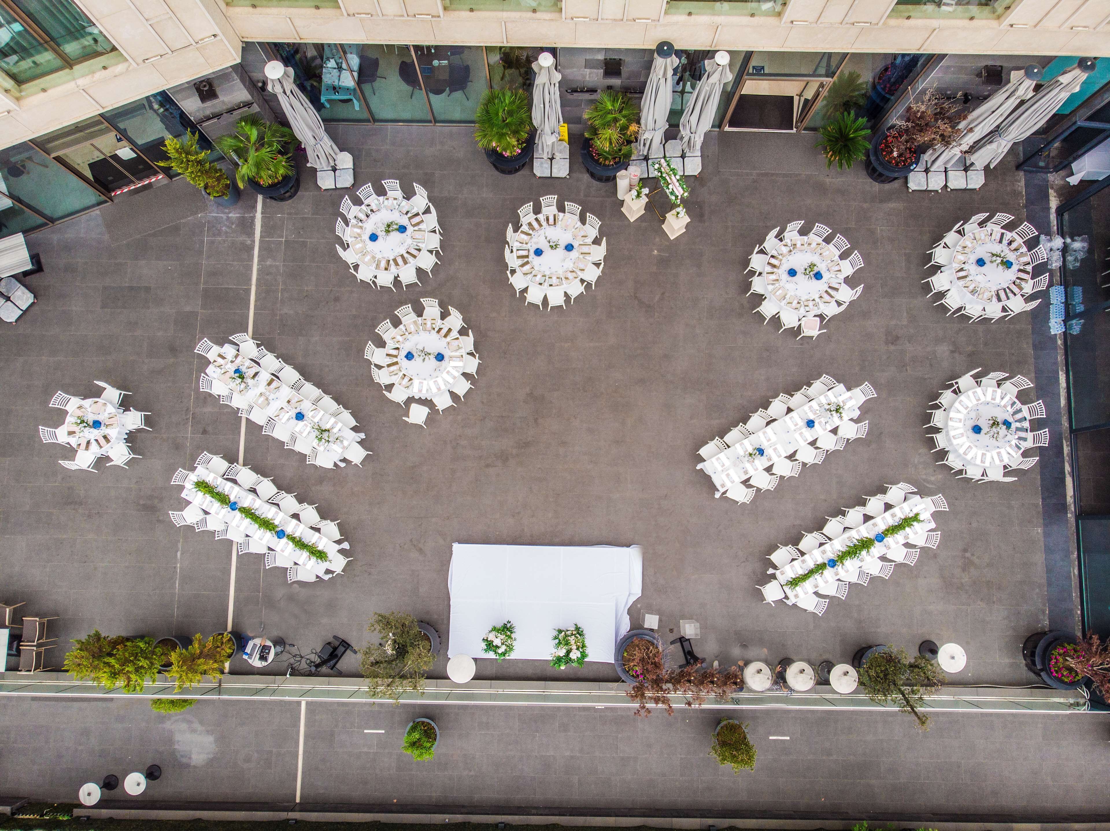 A rooftop terrace with multiple round tables and chairs, surrounded by potted plants and umbrellas.