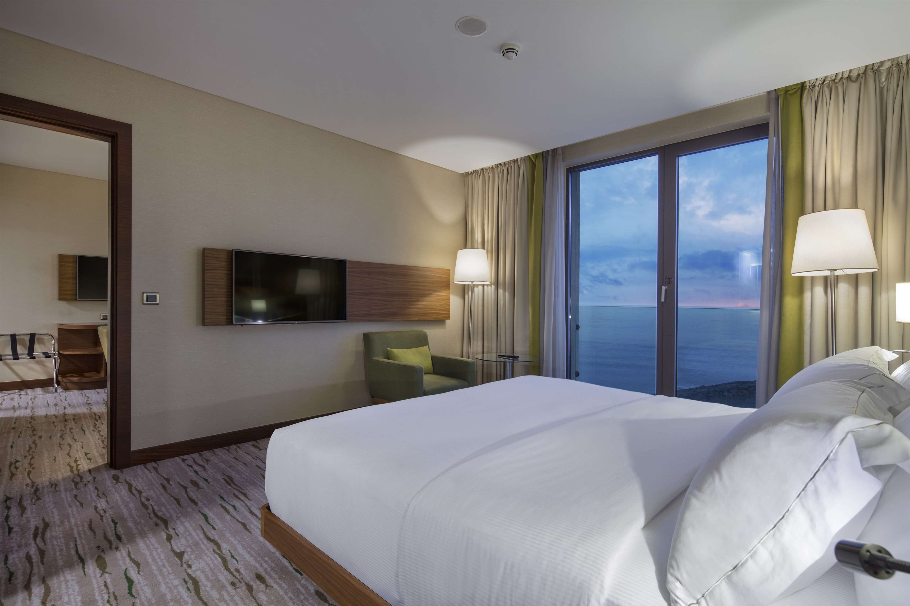 Habitación de hotel con una cama grande, un televisor montado en la pared, una silla y vista al océano a través de un ventanal amplio.