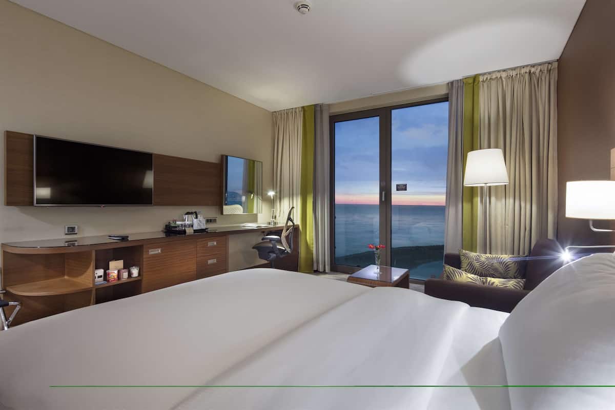 Quarto de hotel com uma cama grande, uma mesa com televisão de tela plana, uma mesinha com abajur e vista para o oceano pelas janelas.