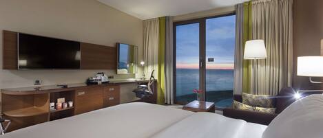 A hotel room with a large bed, a desk with a flat-screen TV, a small table with a lamp, and a view of the ocean through the windows.
