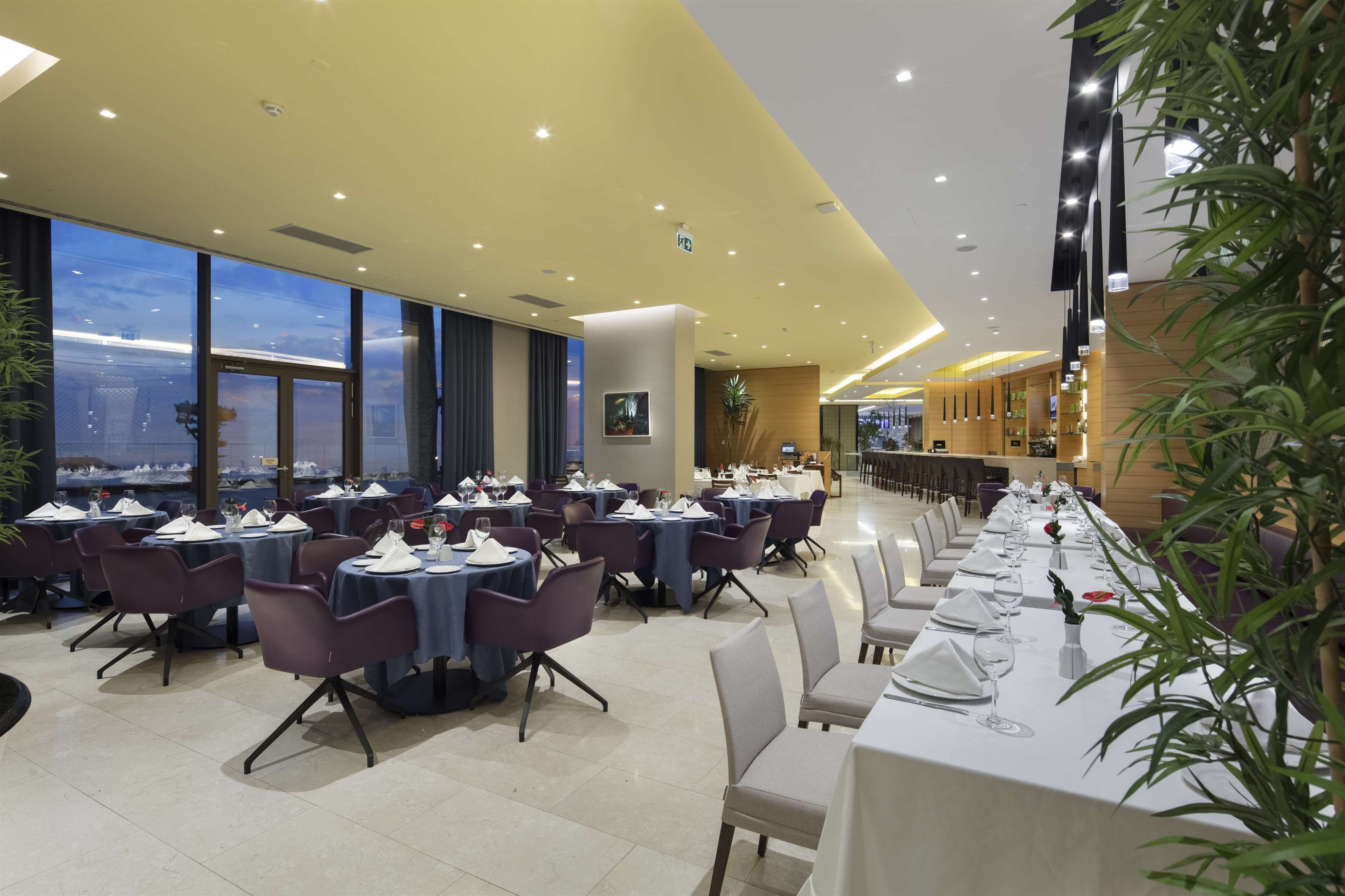 Un restaurante con mesas redondas, sillas moradas y vistas a la ciudad a través de amplios ventanales.
