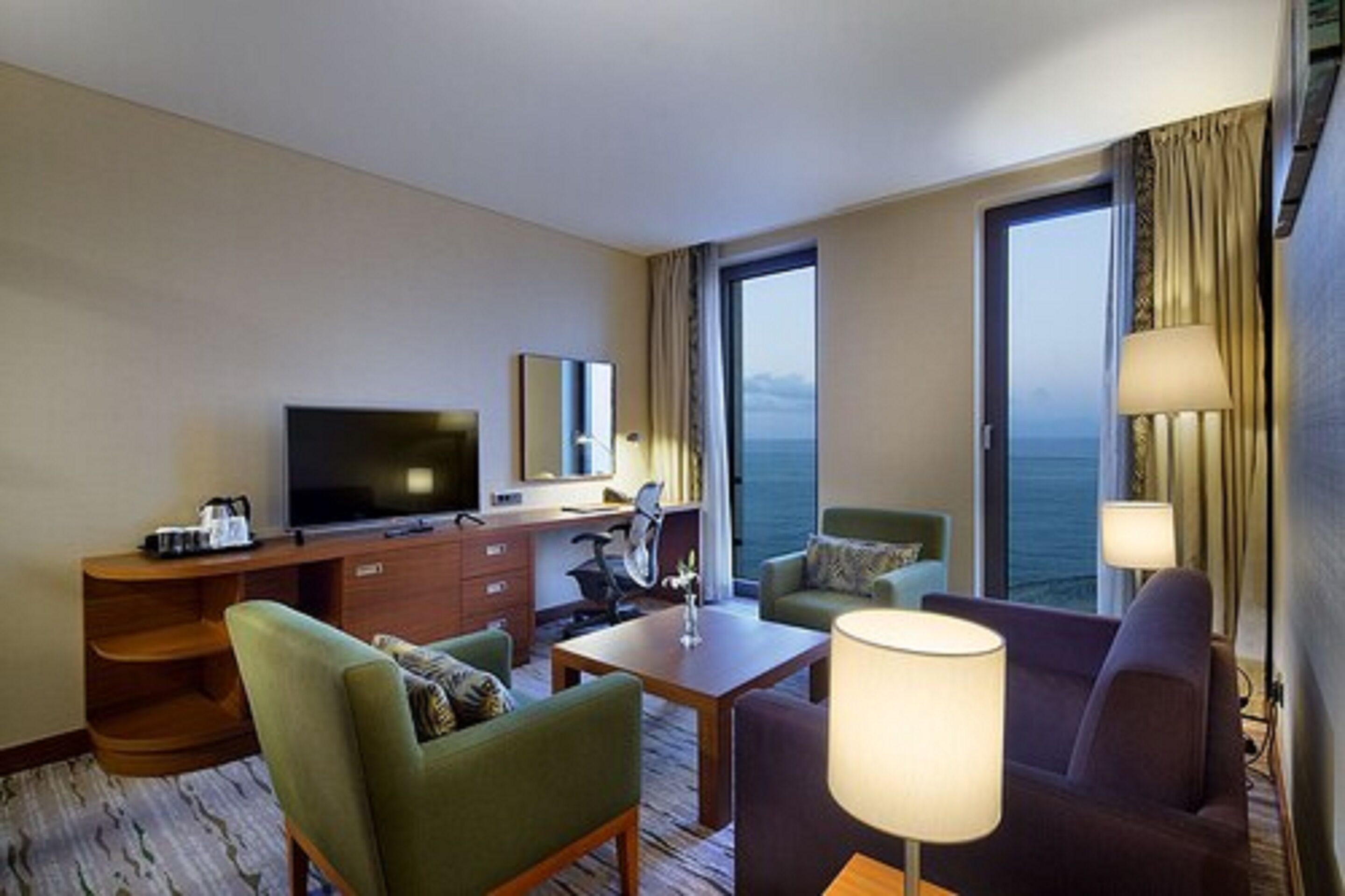 A hotel room with a large TV, a desk, two chairs, a sofa, and a view of the ocean.