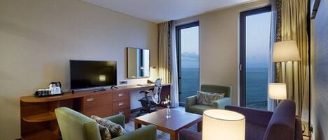 Een hotelkamer met een grote televisie, een bureau, twee stoelen, een bank en uitzicht op zee.