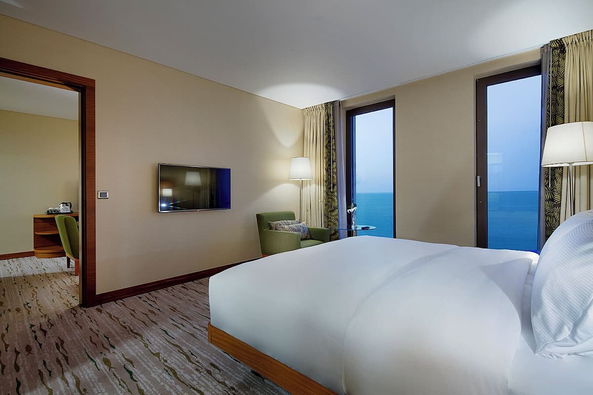Quarto de hotel com uma cama grande, televisão, escrivaninha com cadeira e vista para o mar pelas janelas.