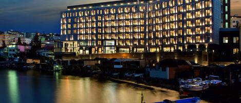 A modern hotel with a large number of uniformly lit windows, situated by a waterfront with boats docked nearby.