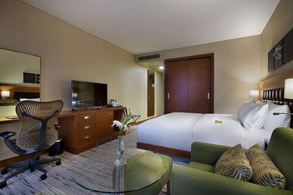 Quarto de hotel com uma cama, uma escrivaninha com cadeira, televisão, penteadeira e uma poltrona verde.