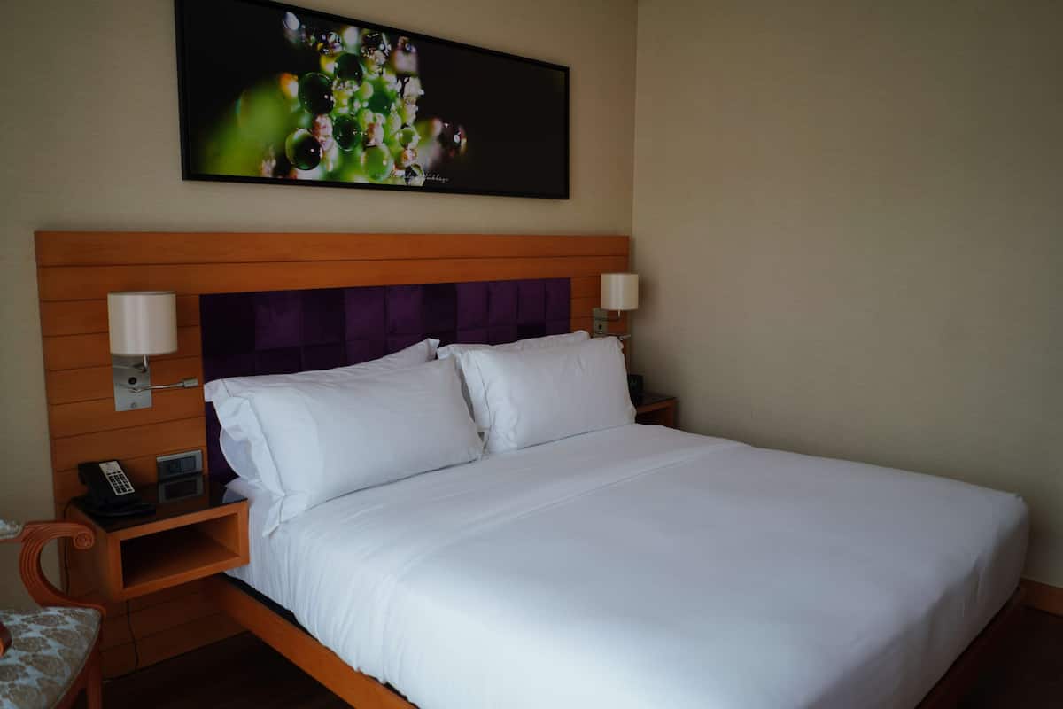 Cama bem arrumada com lençóis brancos, cabeceira de madeira, duas mesas de cabeceira com abajures e uma obra de arte floral sobre a cama.