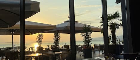 Un restaurante con amplios ventanales que dan a un atardecer, con terraza y macetas.