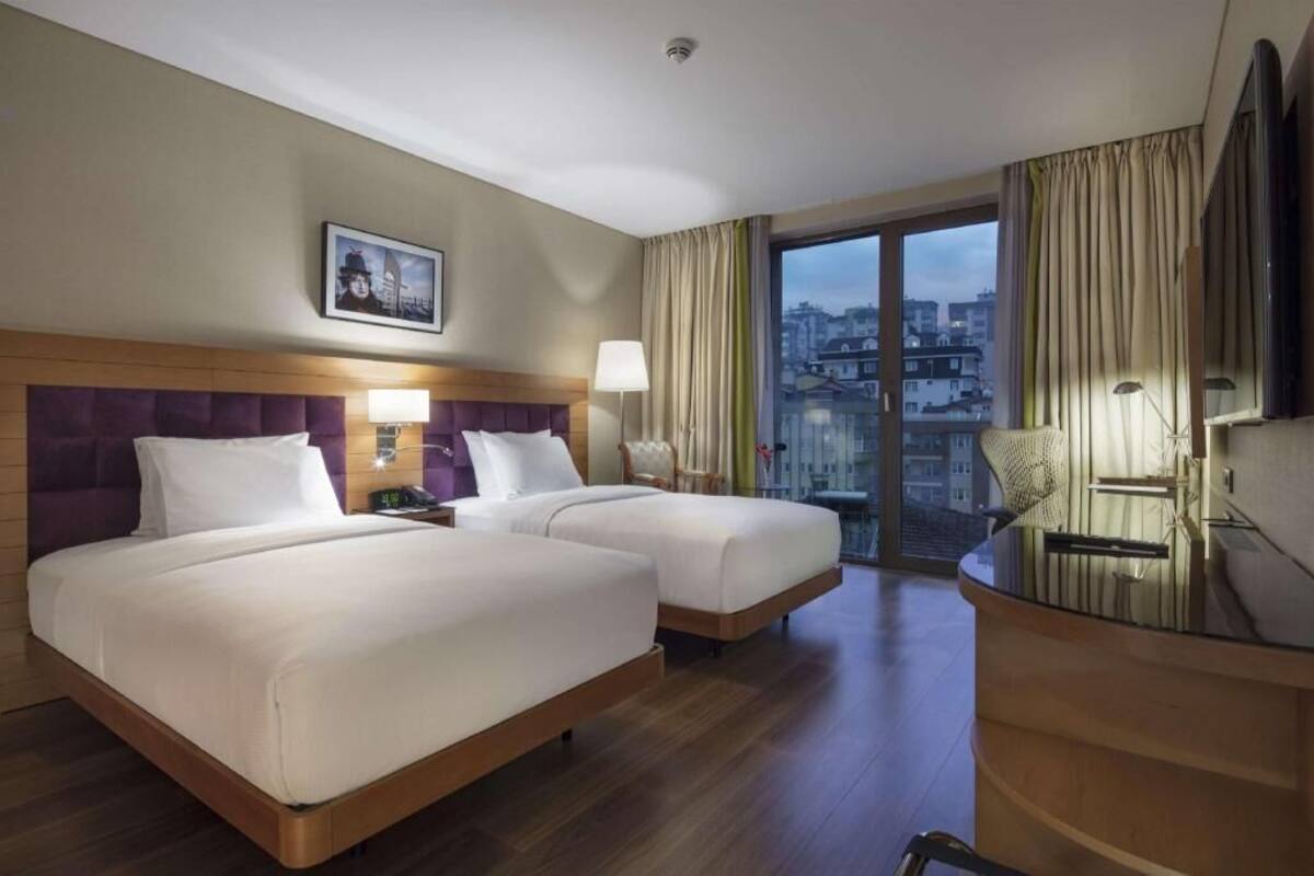 Quarto de hotel com duas camas, uma escrivaninha, uma cadeira, janela com cortinas e vista para a cidade.
