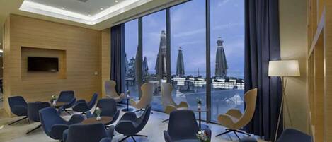 A modern hotel lobby with a view of a cityscape through large windows, contemporary seating, and a flat-screen TV mounted on the wall.