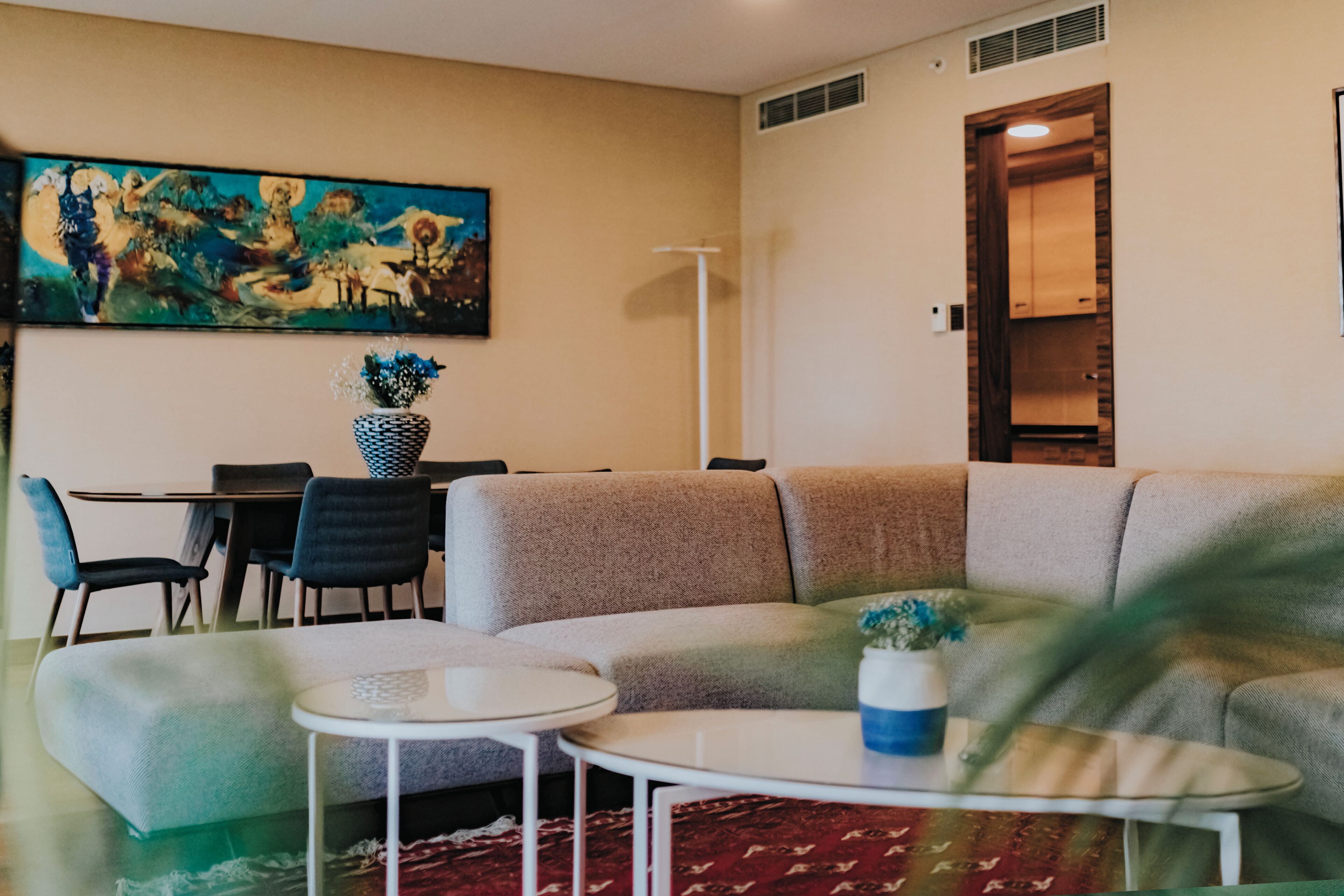 A living room with a sectional sofa, a dining table with chairs, a painting on the wall, and a potted plant.