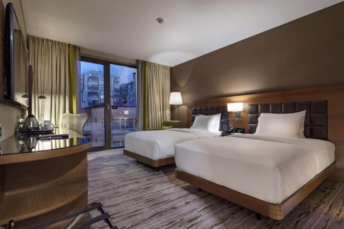 Quarto de hotel com duas camas, uma escrivaninha, uma cadeira, um abajur e vista para a cidade pela janela.