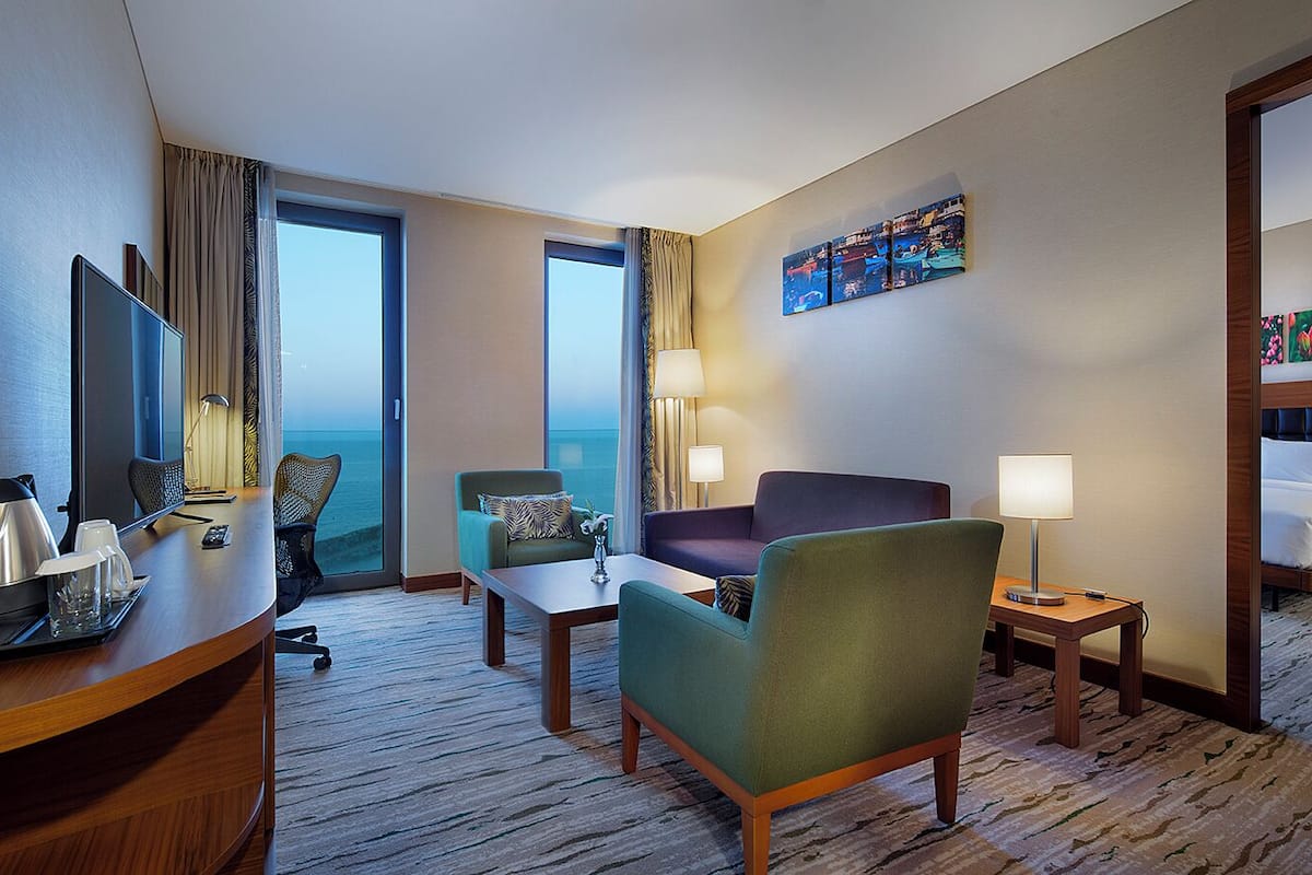 Quarto de hotel com uma escrivaninha, duas poltronas, um sofá, uma cama, uma mesa de cabeceira, um abajur e um quadro na parede.
