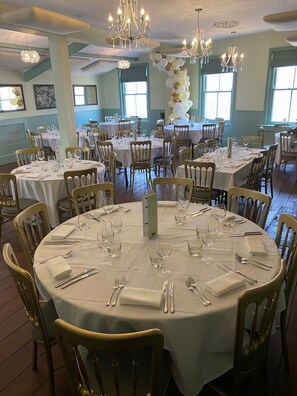 A banquet hall with round tables, chairs, chandeliers, and a balloon arch. - The Peppermill (Devizes)