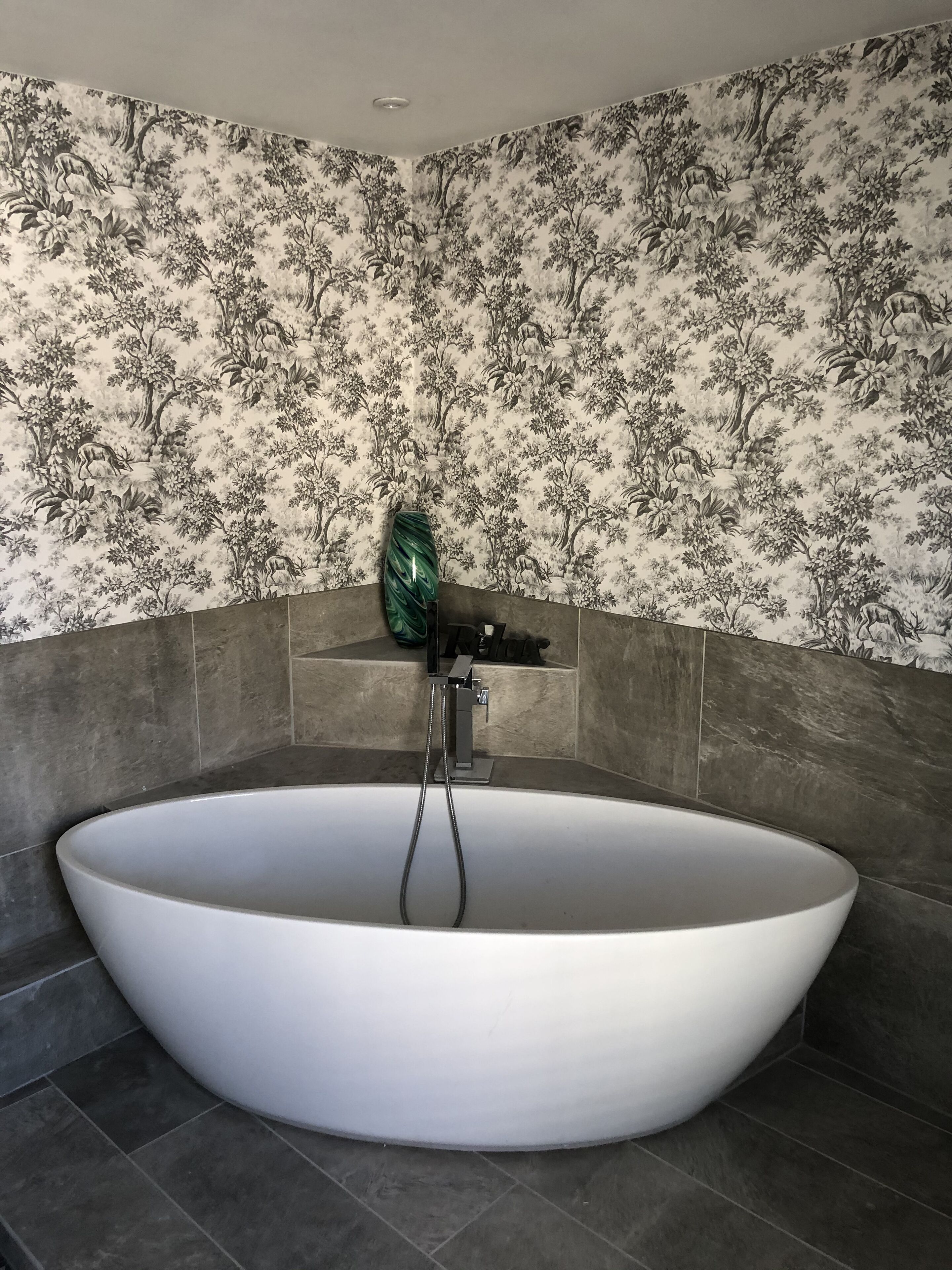 A modern bathroom with a large freestanding bathtub, dark tiled walls, and a floral wallpaper pattern.
