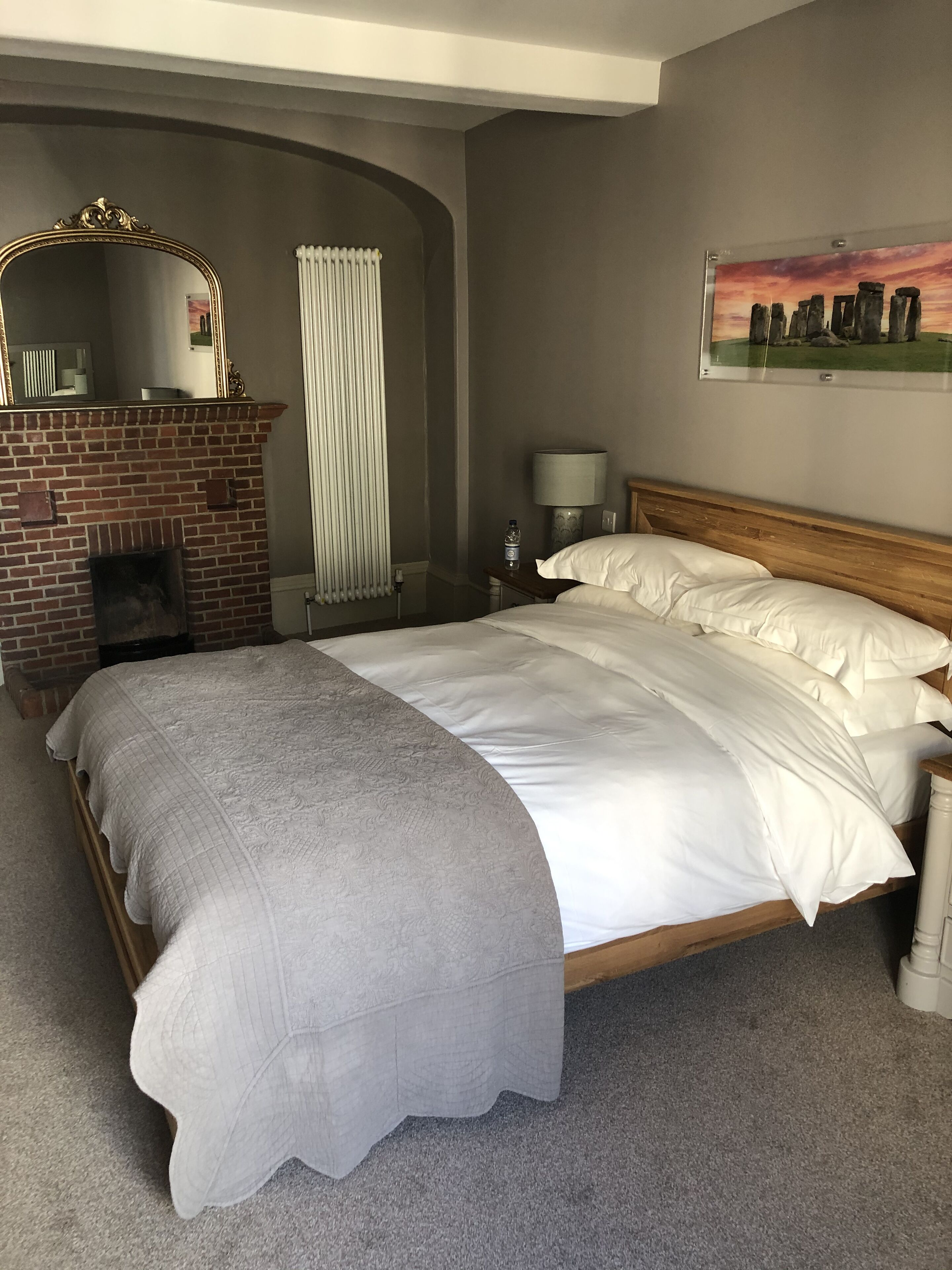 A bedroom with a brick fireplace, a bed with white linens, a nightstand with a lamp, and a painting of Stonehenge on the wall.
